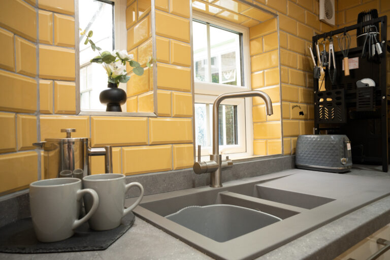 Kitchen sink under the window, with yellow tiles and mugs on the worktop.