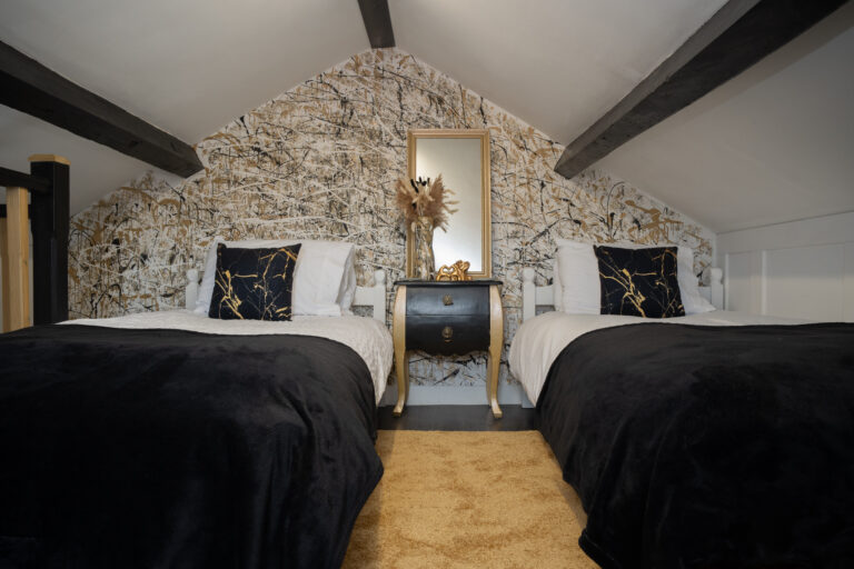Loft bedroom with twin beds either side of a gold mirror, under the beams.