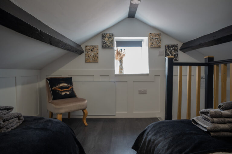 Loft room with a sloping beamed ceiling, a small chair and stairs down.